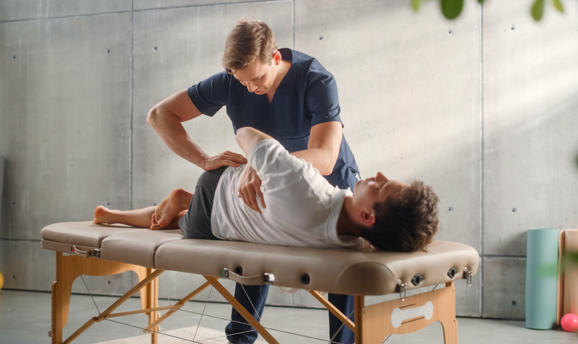 A male chiropractor or physical therapist in dark blue scrubs performing a manual spinal adjustment on a male patient lying on his side on a portable treatment table.